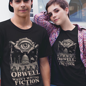 Pode incluir: Uma t-shirt preta com uma estampa gráfica de um olho grande sobre um edifício governamental, câmaras de vigilância e figuras armadas. O texto diz ORWELL WASN'T WRITING FICTION numa fonte estilizada, com um ícone de olho abaixo do texto.