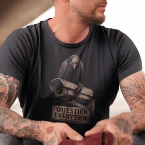 Peut inclure: Un t-shirt noir à manches courtes avec une impression graphique représentant un corbeau sombre perché sur une caméra de surveillance rectangulaire. Sous la caméra, un panneau rectangulaire affiche le texte "QUESTION EVERYTHING" en majuscules.
