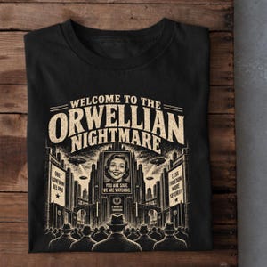 May include: A black t-shirt featuring a beige graphic print with the text WELCOME TO THE ORWELLIAN NIGHTMARE. The design depicts a crowd facing a large screen showing a smiling face with the text YOU ARE SAFE. WE ARE WATCHING. and A BRIGHTER TOMORROW.
