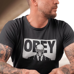 Könnte beinhalten: Ein schwarzes Kurzarm-T-Shirt mit einer weißen rechteckigen Grafik auf der Brust. Die Grafik zeigt das Wort OBEY in fetten schwarzen Großbuchstaben über einer Illustration eines Schädels, der eine Brille und einen Anzug mit Krawatte trägt.