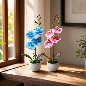 May include: Two artificial orchid plants in small white pots. One plant features vibrant blue flowers with white centers, while the other displays pink flowers with deep magenta centers. Both stems have green leaves and several unopened flower buds.