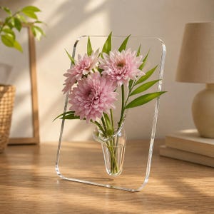 May include: A clear acrylic rectangular frame with a built-in glass bud vase containing three light pink chrysanthemum flowers and green leaves. The transparent structure stands upright, showcasing the floral arrangement within the center.