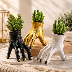 May include: Three hand-shaped planters in black, gold, and white, each featuring a textured wrist with visible stitching details and gold or black painted fingernails. Each planter holds a small green succulent plant in the open palm area.