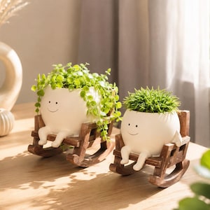 May include: Two white ceramic planters shaped like round characters with simple smiling faces, each sitting in a small brown wooden rocking chair. The planters contain green leafy plants, with one featuring trailing vines and the other a spiky plant.