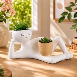 May include: A white ceramic planter shaped like a reclining figure resting on one elbow. The head features a painted face with closed eyes and a small smile, holding a green plant. A separate tan, textured pot sits on the figure's lap.