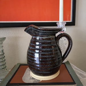 May include: A ceramic pitcher with a dark brown and black mottled glaze featuring horizontal ribbed textures. The vessel has a wide pouring spout, a curved handle, and a smooth, unglazed light beige circular base that sits flat.