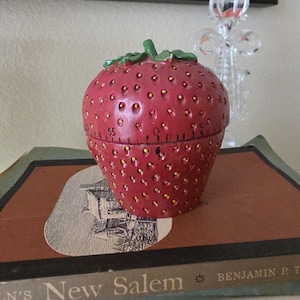 May include: A red strawberry-shaped kitchen timer with a green stem on top. The timer features a textured surface with small yellow dots and black numerical markings around the center, indicating time intervals from zero to fifty-five minutes.