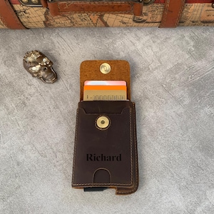 May include: A dark brown leather card holder featuring a snap button closure and the name Richard embossed on the front. The wallet contains several cards with visible numbers and is positioned near a metallic skull-shaped object.