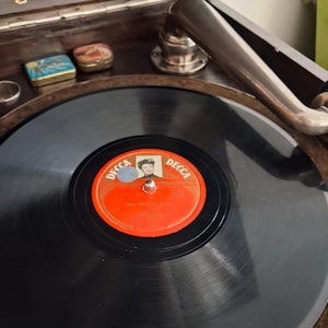 May include: A vintage Columbia Grafonola phonograph featuring a black vinyl record on the turntable. The record has a red Decca label with a portrait and the text Ma Petite Amie. A metallic tone arm rests near the edge of the spinning disc.