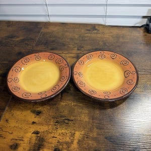 May include: Two round ceramic plates featuring a bright yellow center and a wide, burnt orange rim. The rim is decorated with a repeating pattern of dark brown hand-painted swirls. The edges of the plates are finished with a dark brown border.