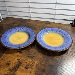 May include: Two round ceramic plates featuring a central yellow circle that transitions into a blue rim. The blue rim is decorated with dark brown hand-painted swirl patterns. Each plate has a dark brown textured edge around the outer circumference.