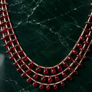 May include: A multi-strand necklace featuring three rows of gold-toned wire strung with numerous small, translucent, deep red teardrop-shaped beads. The beads are evenly spaced along each of the three parallel gold-toned wire strands.