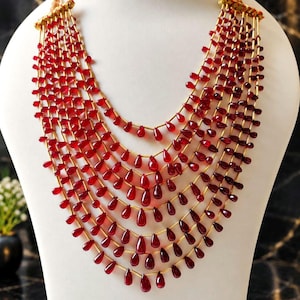 May include: A multi-strand necklace featuring seven cascading layers of deep red, teardrop-shaped beads strung on thin gold-toned wire. The beads are arranged in a graduated pattern, creating a full, layered look that drapes in a wide, curved shape.