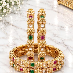 Può includere: Un paio di bracciali rigidi color oro con un design in metallo testurizzato e perlato. I bracciali sono decorati con pietre quadrate rosse e verdi alternate, pietre bianche trasparenti e piccole perle bianche in un motivo ripetuto.