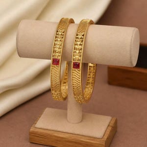 Peut inclure: Deux bracelets rigides dorés ornés de motifs floraux en relief complexes et de textures en lignes ondulées. Chaque bracelet est centré par une pierre précieuse rouge de taille carrée. La surface métallique présente une finition mate.