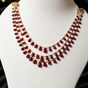 May include: A multi-strand necklace featuring three rows of deep red, teardrop-shaped beads strung on thin gold-toned wire. The necklace is secured at both ends with gold-toned metal clasps, creating a layered, cascading design.