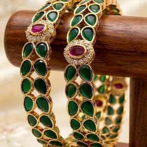 Peut inclure: Deux bracelets rigides en or ornés d'un motif répétitif de pierres vertes en forme de goutte. Chaque bracelet est agrémenté d'une pierre rose ovale centrale entourée de petits cristaux clairs, créant un bijou décoratif et symétrique.