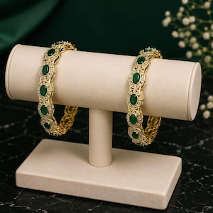 May include: Two gold-toned bangles featuring a series of oval-shaped green stones surrounded by small clear crystals. The bracelets are displayed on a beige T-bar stand, showcasing a repeating pattern of emerald-colored gems and metallic accents.