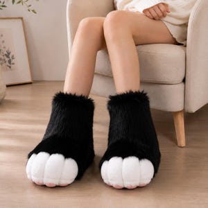 Op de afbeelding: Een paar enkelhoge pantoffels met een zwarte imitatiebont buitenkant en witte pluche teenkussentjes aan de voorzijde. De pantoffels zijn ontworpen als dierenpoten, met zachte, afgeronde witte delen bij de tenen en een dikke, donkere harige textuur.