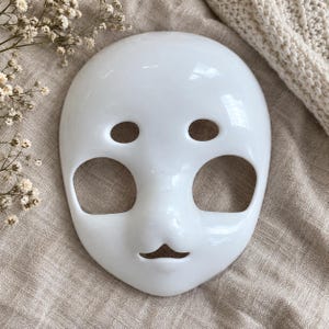 Op de afbeelding: Een glad, glanzend wit gezichtsmasker met een afgeronde vorm, voorzien van twee ovale oogopeningen, twee grote ronde wanguitsnijdingen en een kleine driehoekige mondopening, inclusief een licht gevormd en uitstekend neusgedeelte.