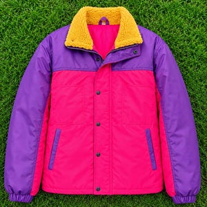 Op de afbeelding: Een color-block jas met een felroze middenpaneel en mouwen, gecombineerd met een contrasterend paars gedeelte op de borst en schouders. De jas is voorzien van een textuurrijke mosterdgele sherpa kraag, een ritssluiting en drukknopen.