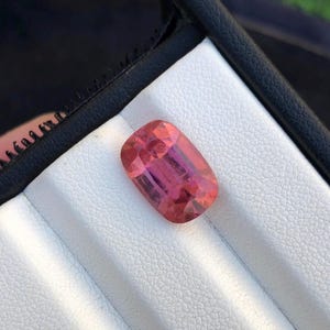 Puede incluir: Una piedra preciosa de corte cojín con un tono rosa y rojizo vibrante. La piedra presenta una superficie facetada que refleja la luz, mostrando claridad interna y saturación de color. Está colocada sobre una almohadilla de exhibición blanca.