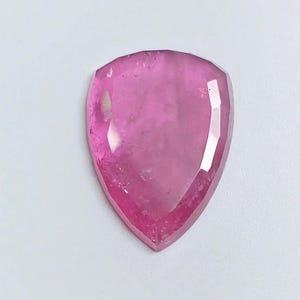 May include: A pear-shaped, faceted gemstone in a vibrant, translucent pink hue. The stone features a flat, polished surface with multiple geometric facets along the edges, creating a distinct shield-like outline with a pointed bottom tip.