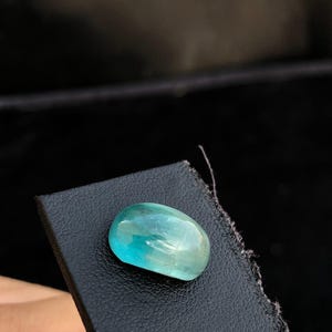 May include: A polished, oval-shaped gemstone with a translucent, vibrant teal-blue color. The stone features a smooth, rounded cabochon cut that reflects light across its surface, displaying subtle internal variations and a clear, glassy texture.