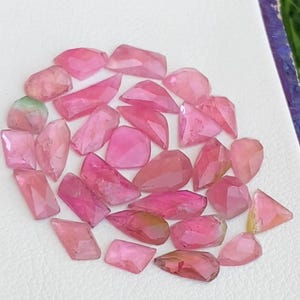 May include: A collection of loose, faceted pink gemstones in various shapes and sizes. The stones exhibit translucent qualities with varying shades of light to deep pink, some featuring subtle green undertones, arranged on a white textured surface.