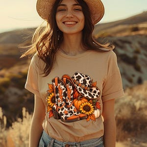 May include: A beige short-sleeved t-shirt featuring a graphic print of a pair of cow-print cowboy boots, a matching cow-print cowboy hat, bright yellow sunflowers, and orange pumpkins, all arranged in a cluster on the front of the garment.
