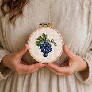 Peut inclure: Un tambour à broder circulaire en bois contenant un motif au point de croix représentant une grappe de raisins violets avec une feuille et une vrille vertes. Le tissu est de couleur crème et le tambour possède une vis de serrage en laiton.