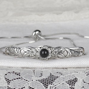 May include: A silver-toned bracelet with an adjustable chain. The bracelet features a central floral design with a black gemstone. The decorative band is flanked by intricate floral patterns. The bracelet rests on a white surface with lace trim.
