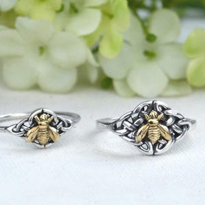 May include: Two silver rings with a gold-colored bee design. The bees are centered on a decorative silver setting. The rings are displayed in front of a blurred background of white and pink flowers.