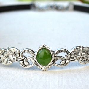 May include: A silver headband with a green gemstone and floral design. The headband is made of a black leather band.