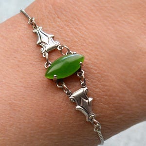 Puede incluir: Un brazalete de plata con una gema verde ovalada en el centro. El brazalete tiene eslabones decorativos de plata a cada lado de la gema y una fina cadena. El brazalete se muestra en una muñeca. La gema mide aproximadamente 1,5 cm.