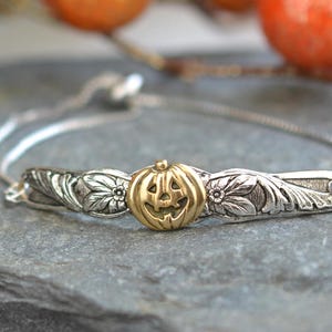 May include: Silver-toned adjustable bracelet with a gold-colored pumpkin charm. The bracelet features a detailed floral design on the sides of the pumpkin. The background includes blurred orange and brown decorative spheres, suggesting a fall or Halloween theme.