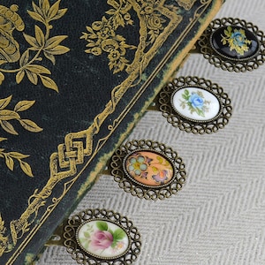 May include: Five ornate metal bookmarks with floral designs on porcelain inserts. The bookmarks are attached to a vintage book with a gold and black cover.