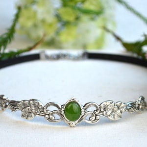 May include: A silver headband with a green gemstone and floral design. The headband is made of a black elastic band.