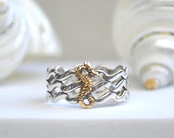 May include: A silver ring with a gold seahorse design, set against a backdrop of two white seashells. The ring features a wavy pattern and is a nautical-themed jewelry piece.
