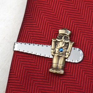 May include: A red herringbone patterned tie with a silver tie clip featuring a bronze-colored nutcracker figure with a blue gem. The tie clip is approximately 2 inches long.