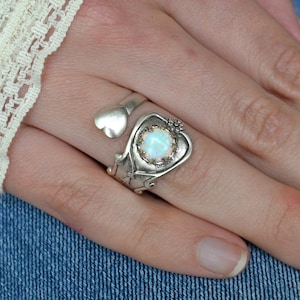 May include: A silver ring with a heart-shaped design and a white opal gemstone. The ring is set on a finger with a white lace background.
