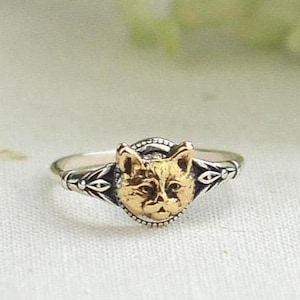 May include: A silver ring with a gold cat face design. The cat face is centered on the ring and is surrounded by a decorative border. The ring has a delicate, detailed design.