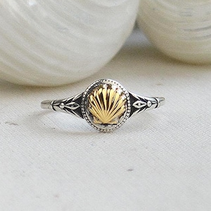 May include: A silver ring with a gold seashell design is displayed in front of two large, white seashells. The ring has a detailed band and a gold-colored seashell center, with a small, clear stone border.