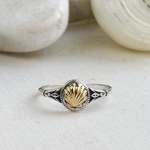 May include: A silver ring with a gold seashell design. The ring has an oval-shaped gold center with a detailed seashell pattern, framed by a silver band with decorative accents. The ring is displayed on a white surface.