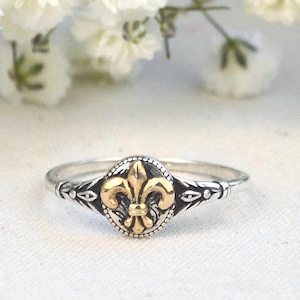 May include: A silver ring with a gold fleur-de-lis design at the center. The ring has a detailed band with small decorative elements. White flowers are in the background, creating a soft, elegant setting.