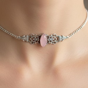 May include: A silver choker necklace with an oval, light pink gemstone centerpiece. The necklace features an ornate, filigree design around the gemstone and a delicate chain. The jewelry is displayed on a person's neck.