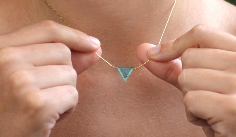 May include: A delicate gold necklace featuring a triangular, light blue gemstone pendant. The pendant is suspended from a thin gold chain. The necklace is being held up by two hands.