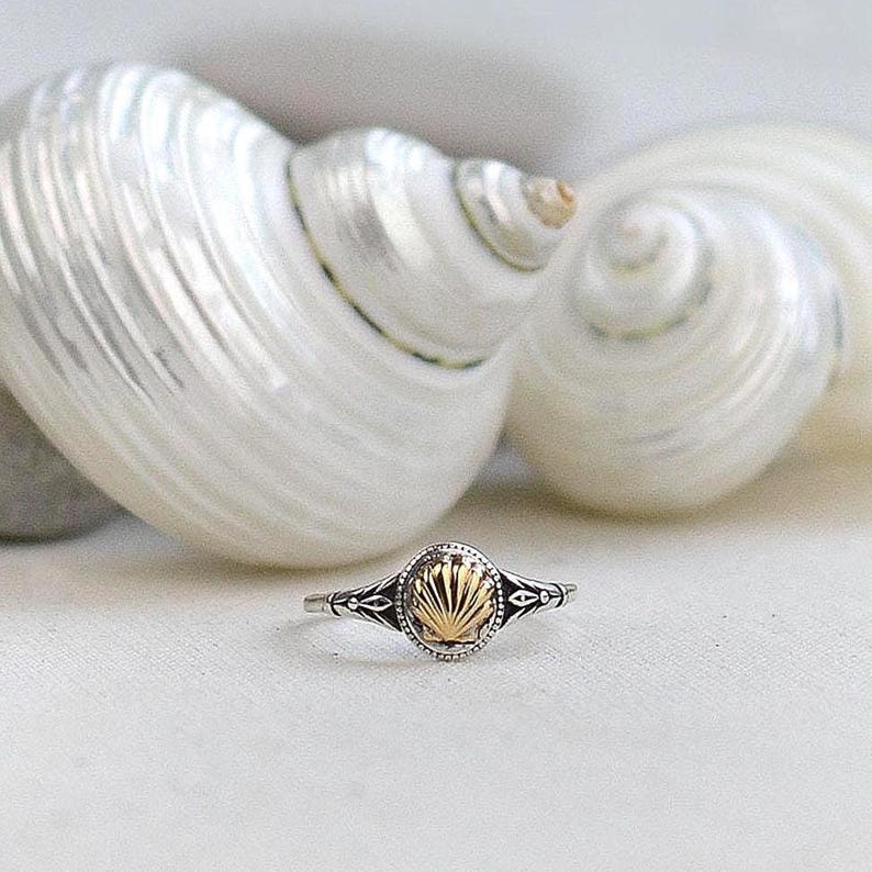 May include: A silver ring with a gold seashell design. The ring features an oval-shaped gold seashell set in a silver band with intricate detailing. The ring is set against a white background with two large, white seashells.