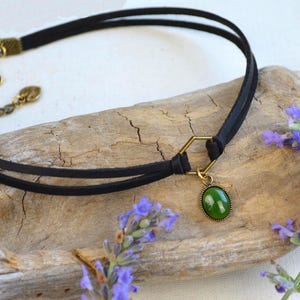 May include: A black choker necklace with a double-strand design. It features a gold-colored clasp and a hexagonal connector holding a green oval pendant. Lavender sprigs with purple flowers are in the background.