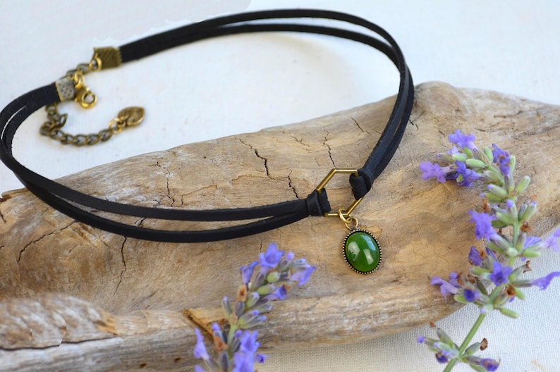 May include: A black choker necklace with a double-strand design. It features a gold-colored clasp and a hexagonal connector holding a green oval pendant. Lavender sprigs with purple flowers are in the background.